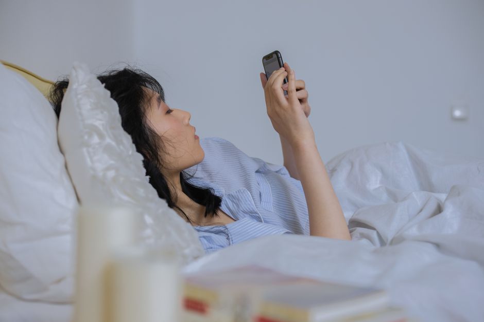 Asian woman lying in bed at home using smartphone, in a cozy setting.