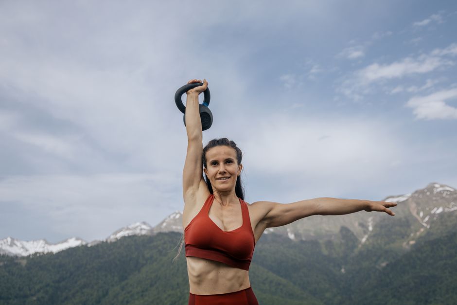 Woman in activewear lifting a kettlebell outdoors with mountain scenery, showcasing strength and fitness.