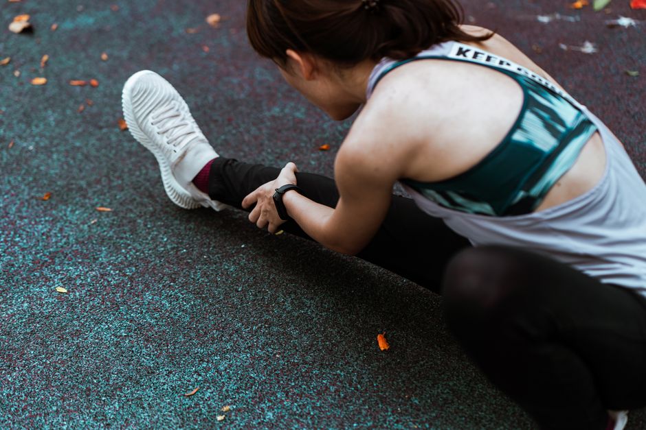 From above side view of crop anonymous female athlete in sportswear leaning forward while warming up on asphalt embankment with faded leaves in fall