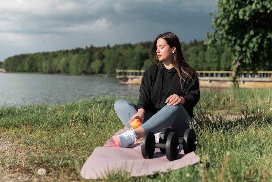 Woman sitting on yoga mat beside a lake with exercise equipment, enjoying a calm outdoor workout.