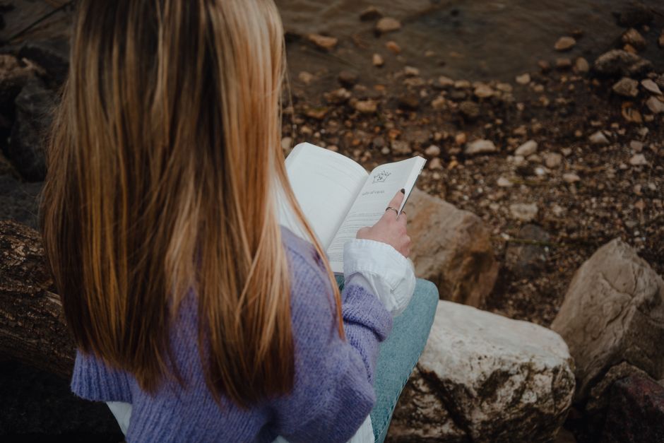 Peaceful scene of a woman reading a book by the riverside, showcasing relaxation in nature.