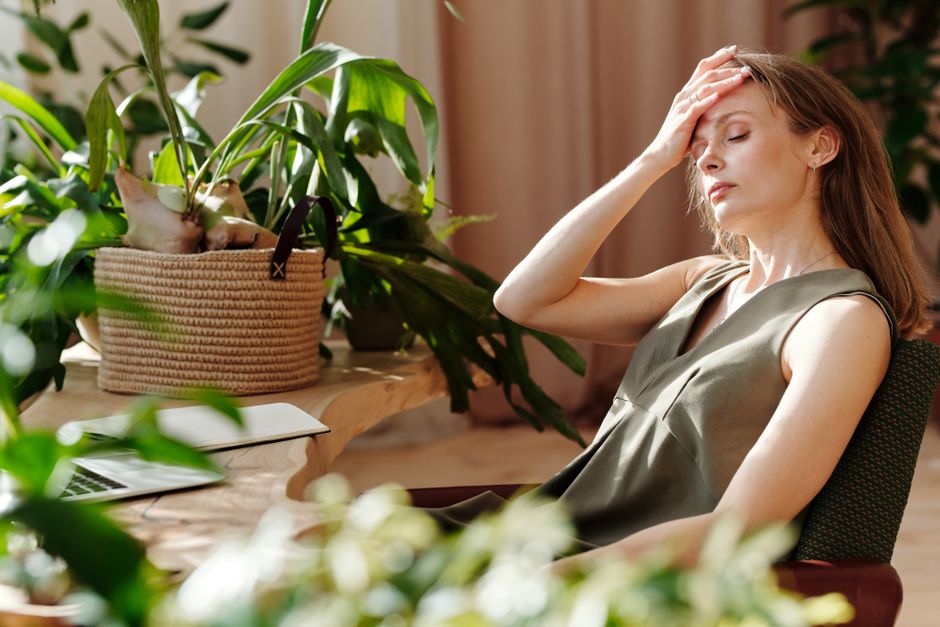 Woman with hand on forehead sitting indoors among plants, showing stress or fatigue.