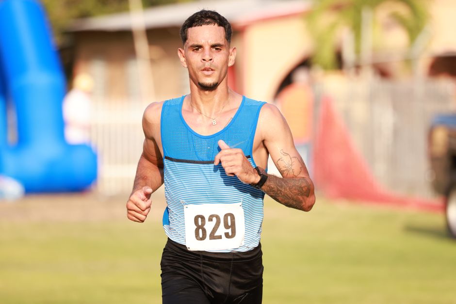 Male athlete running in a marathon under bright sunlight, wearing a blue vest and displaying focus.