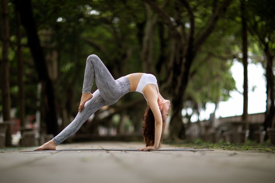 A woman performs a yoga pose outdoors, demonstrating flexibility on a tree-lined walkway.