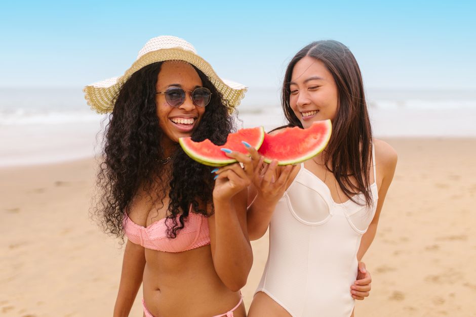 Two women in swimsuits share watermelon on a sunny beach, embodying summer fun.