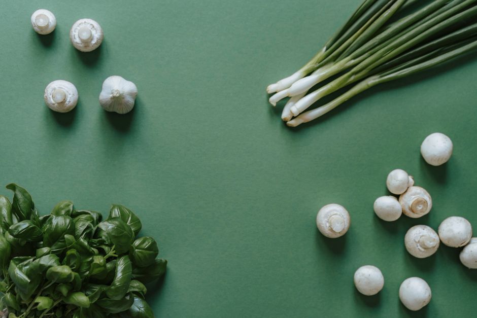 Top-down view of fresh ingredients including basil, mushrooms, garlic, and green onions on a green background.