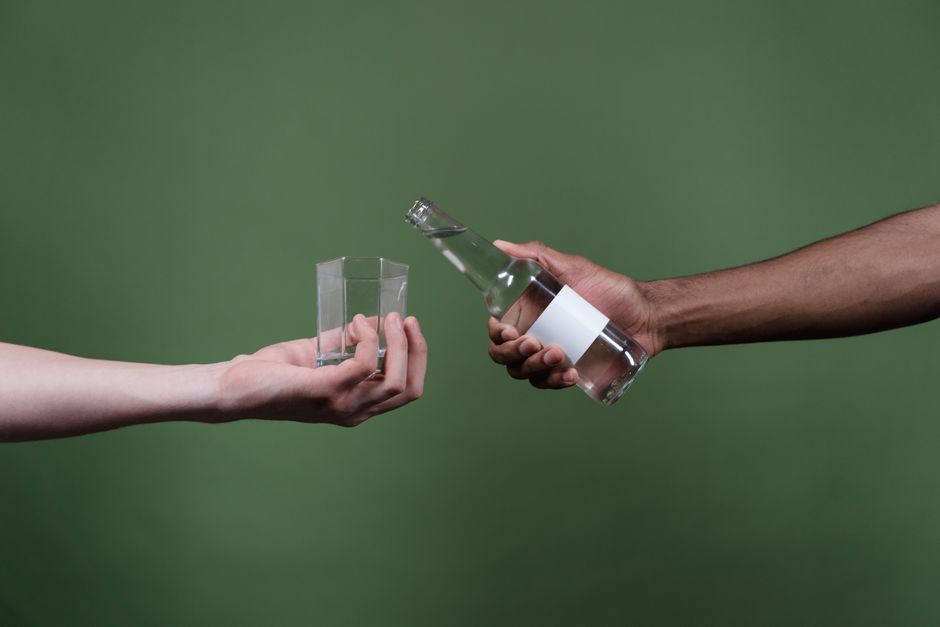 Close-up of diverse hands holding a clear bottle and glass, set against a green background.