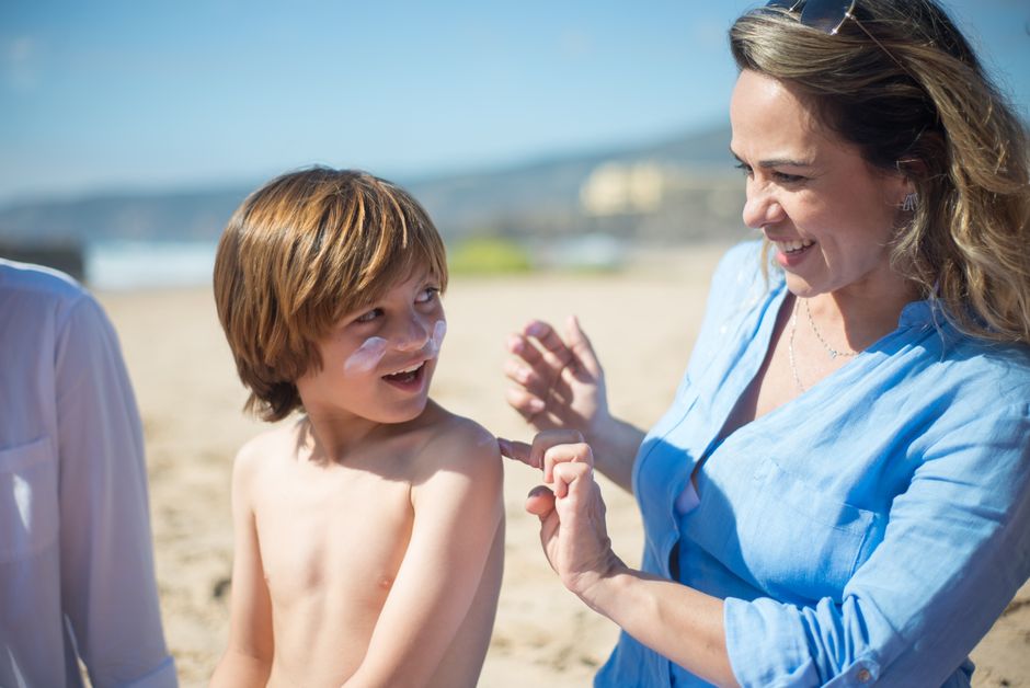 A woman applying sunscreen to her smiling son on a sunny beach day, promoting skin care and safety.