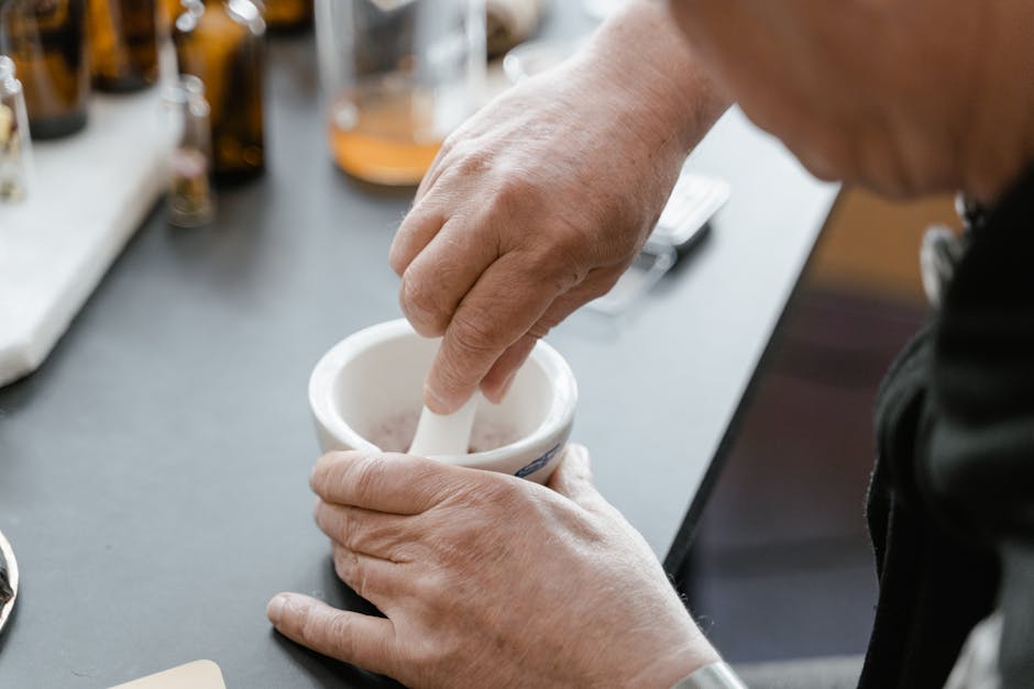 Hands using a mortar and pestle in a lab setting with glass bottles in the background.