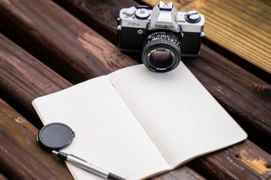 A vintage Minolta camera beside an open notebook and pen on a wooden surface.