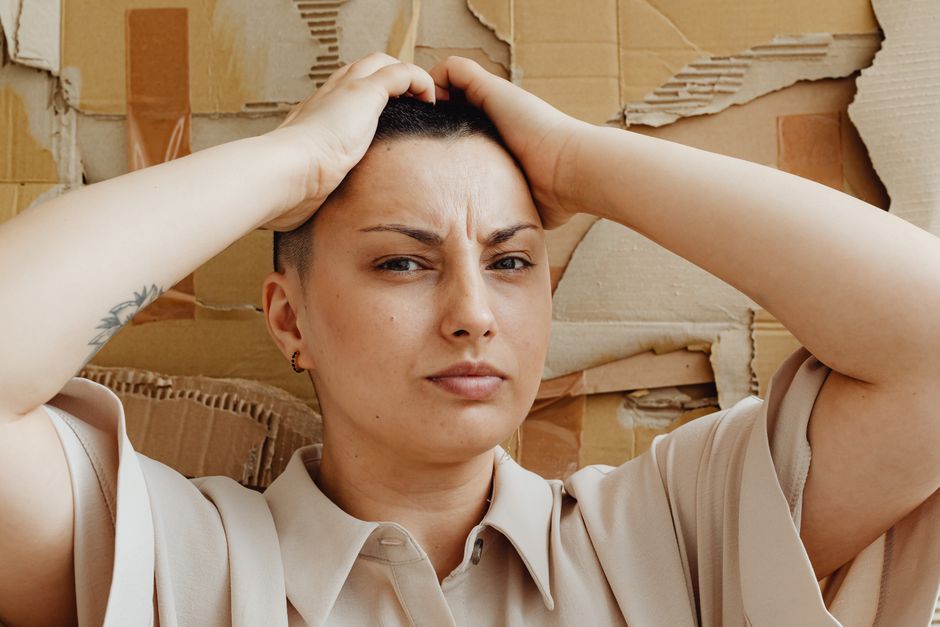 A close-up view of a woman with short hair, hands on head, indoors with a cardboard background.