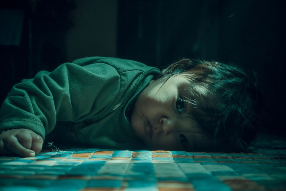 A serene, moody portrait of a child lying on a vibrant patterned floor indoors.