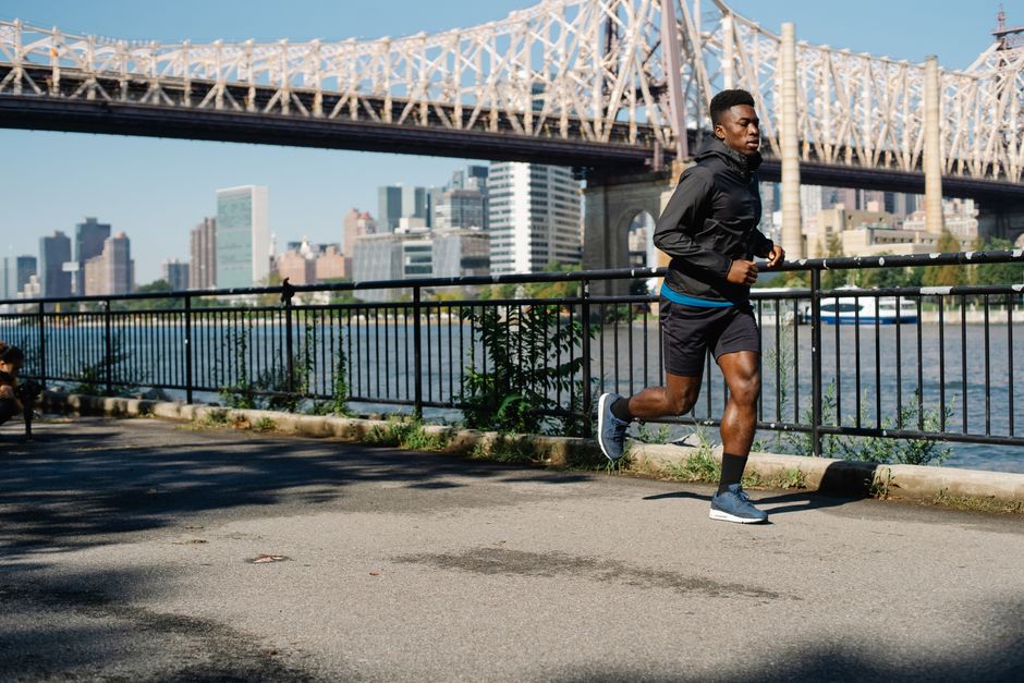 African American man jogging by the river with an urban bridge and city skyline in the background, promoting healthy living.
