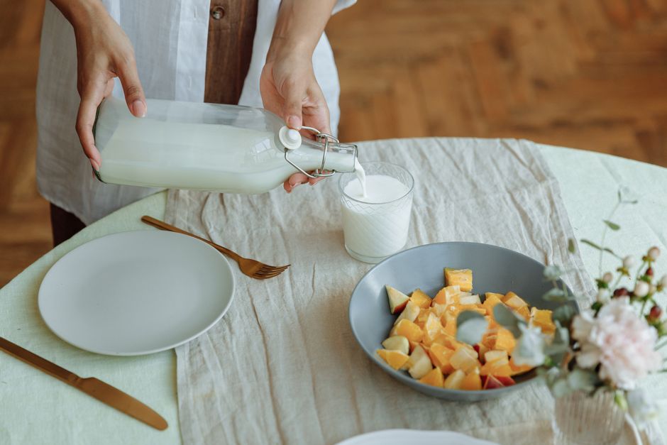 Close-up of hands pouring milk into glass alongside fruit salad, set for breakfast.