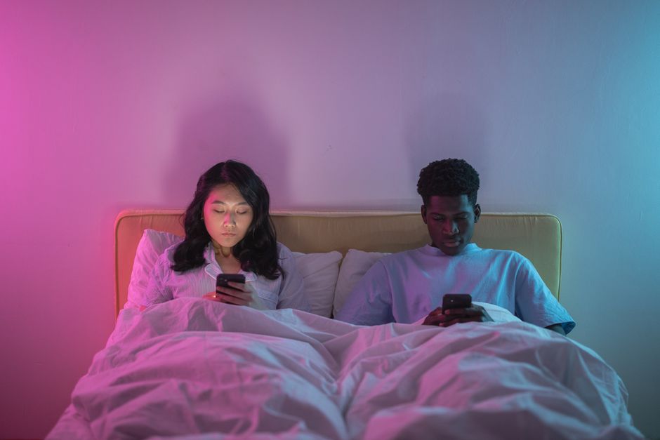 Couple lying in bed using smartphones under colored lights, showcasing modern digital lifestyle.