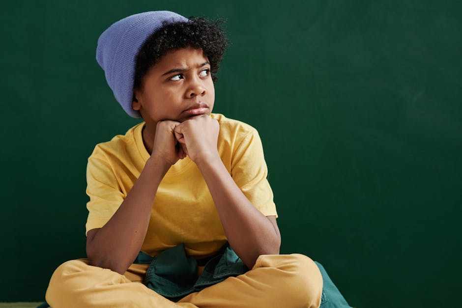 Pensive boy with curly hair wearing a yellow shirt and blue beanie against a green background.