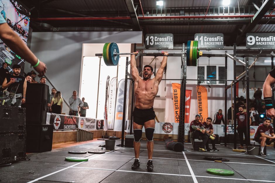 A muscular athlete performs a powerful overhead lift during a CrossFit competition indoors, showcasing strength and determination.