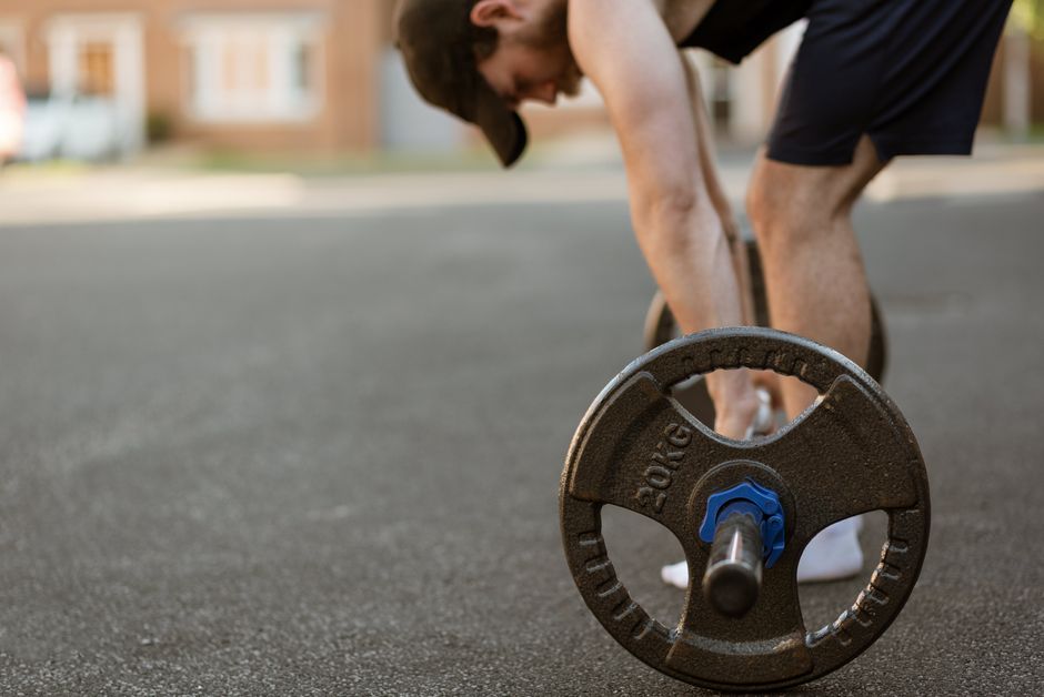 Side view of crop young bearded bodybuilder leaning forward with heavy barbell on asphalt road in town