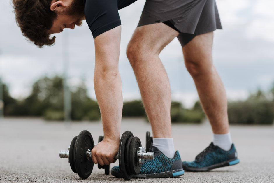 Side view of crop sporty male lifting massive metal dumbbell on asphalt road on blurred background of green trees