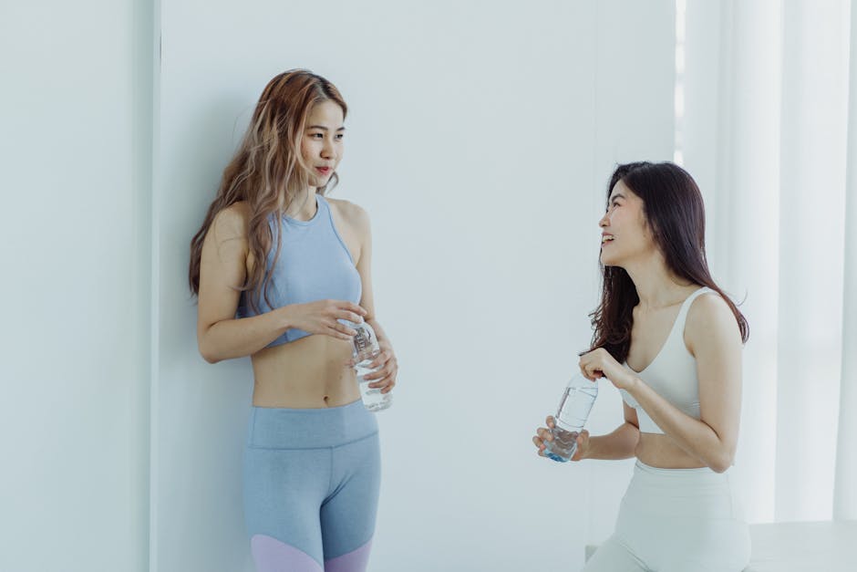 Two women in activewear enjoying a casual conversation while holding water bottles indoors.