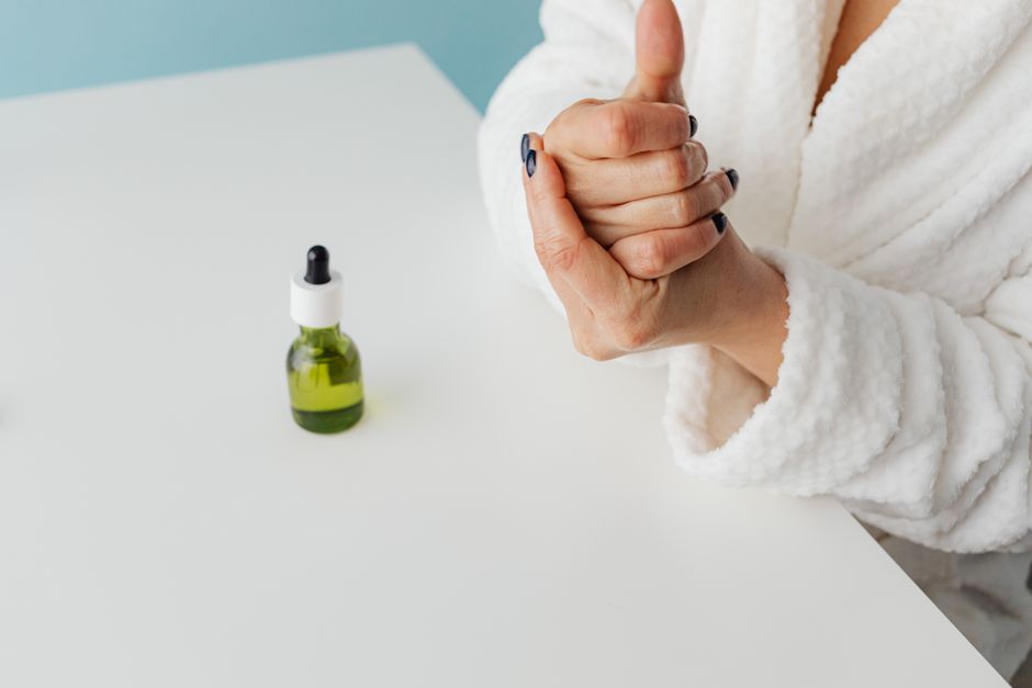 Close-up of a woman in a robe applying moisturizing serum to her hands with a dropper bottle nearby.